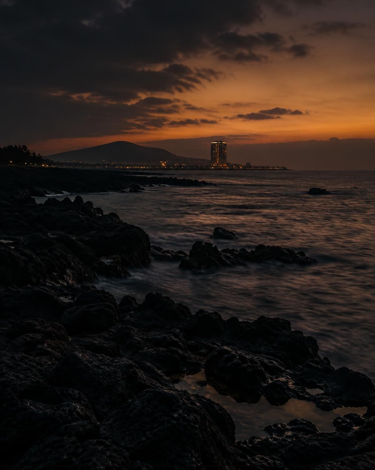 黄昏时济州海岸的黑色火山岩，远处是亮灯的酒店高楼